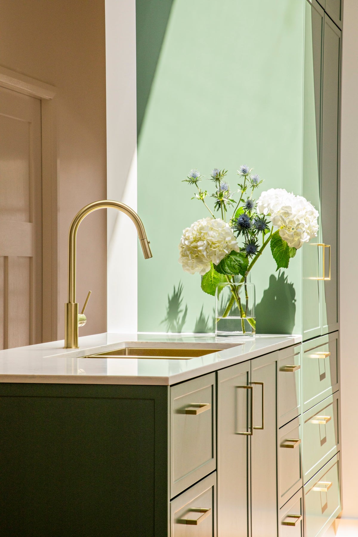 Brass kitchen mixer above a white sink with sage green cabinetry and flowers